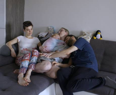 dvě ženy a jeden muž se choulí spolu na gauči. prostřední žena drží dítě v šátku